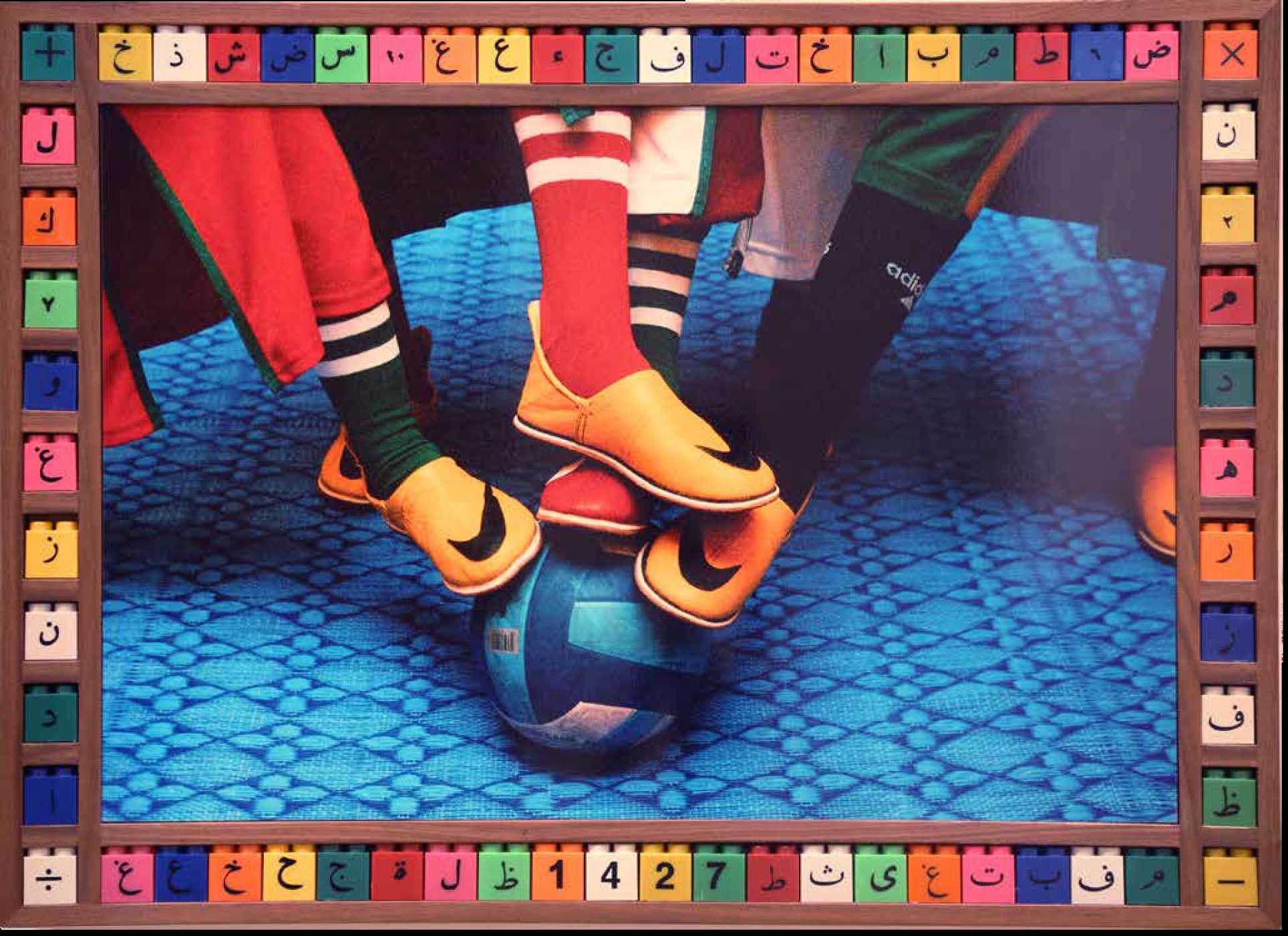 Titled ‘Feet Ball’ by Haddan Hajjaj. 2006. Metallic Lambda print on wood frame with objects, 62X82cm. In this photograph, four feet wearing brightly colored slippers jostle for control of a football. There is a ‘swoosh’ logo of a universally recognized sports brand. This inventive fusion combines local tradition with a commentary on the power of branding in global capitalism. The piece is framed by a mosaic of 62 colored plastic children’s building blocks printed with Arabic letters, numbers and mathematical symbols.